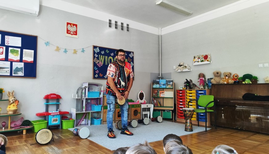 Mężczyzna w kolorowej koszuli gra na bębnie djembe przed grupą dzieci w sali przedszkolnej.