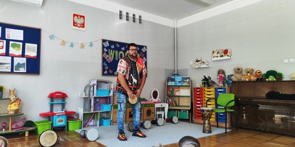 Mężczyzna w kolorowej koszuli gra na bębnie djembe przed grupą dzieci w sali przedszkolnej.