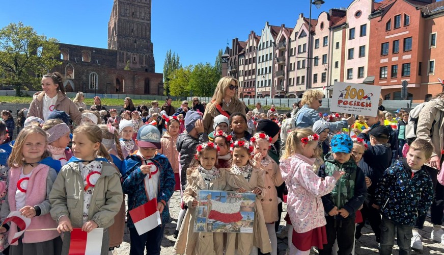 Dzieci z polskimi flagami i wiankami na głowach stoją na placu w Gdańsku. W tle gotyckie ruiny i kolorowe kamienice.