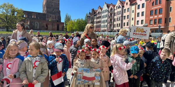 Dzieci z polskimi flagami i wiankami na głowach stoją na placu w Gdańsku. W tle gotyckie ruiny i kolorowe kamienice.