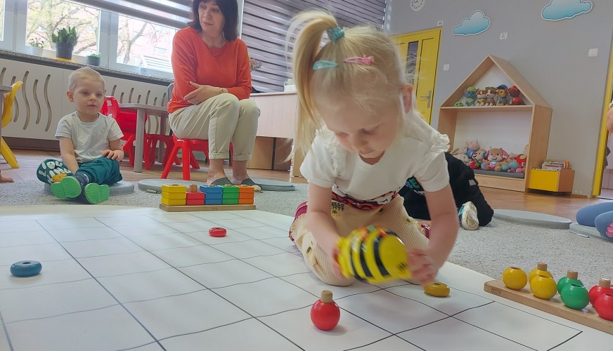 Dzieci siedzą na podłodze w sali przedszkolnej i układają kolorowe zabawki na macie z siatką. W tle nauczycielka obserwuje.